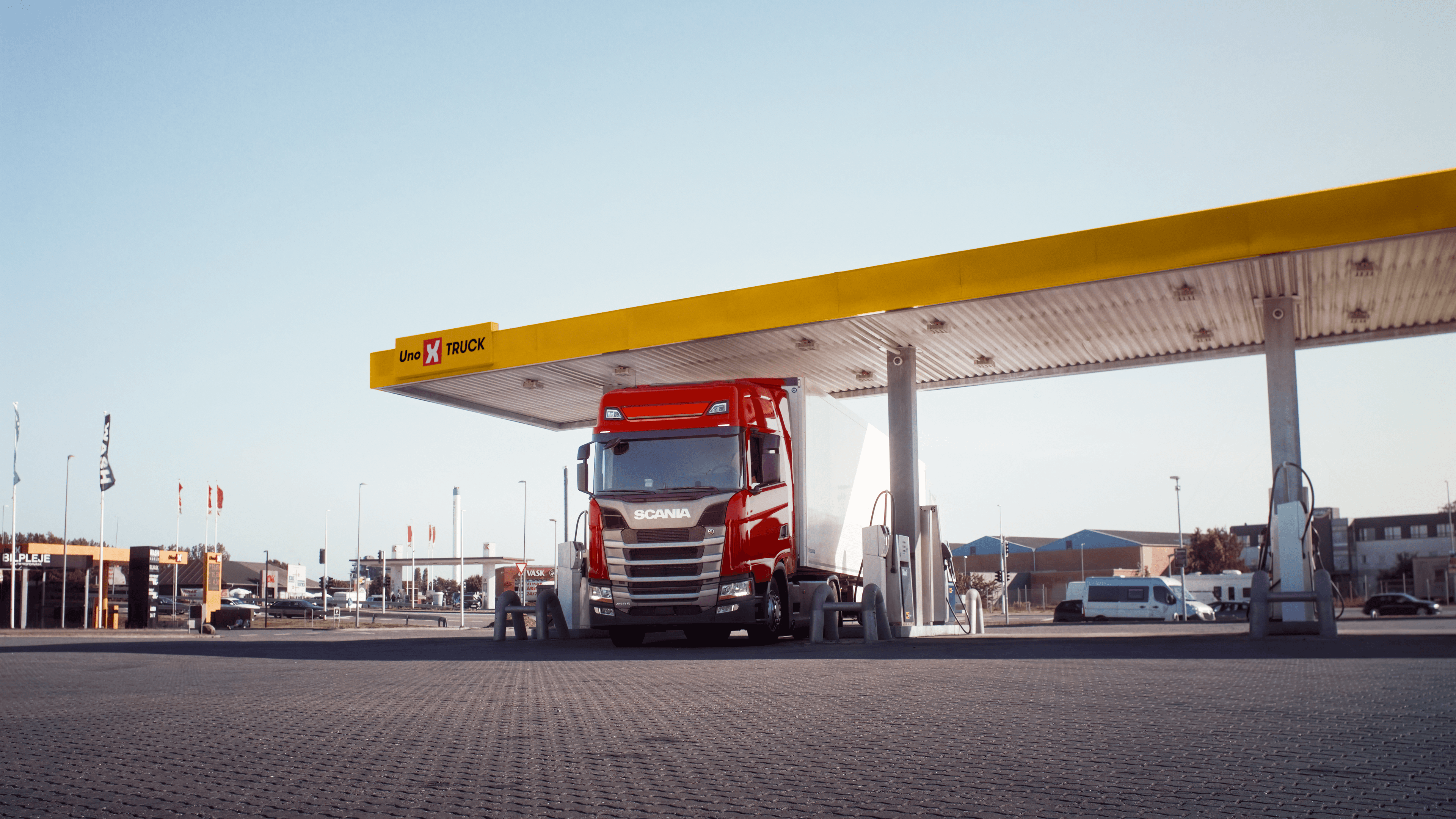 Rød Scania lastbil ved Uno-X Truck station, klar til tankning under gul overdækning, med bygninger og flag i baggrunden.