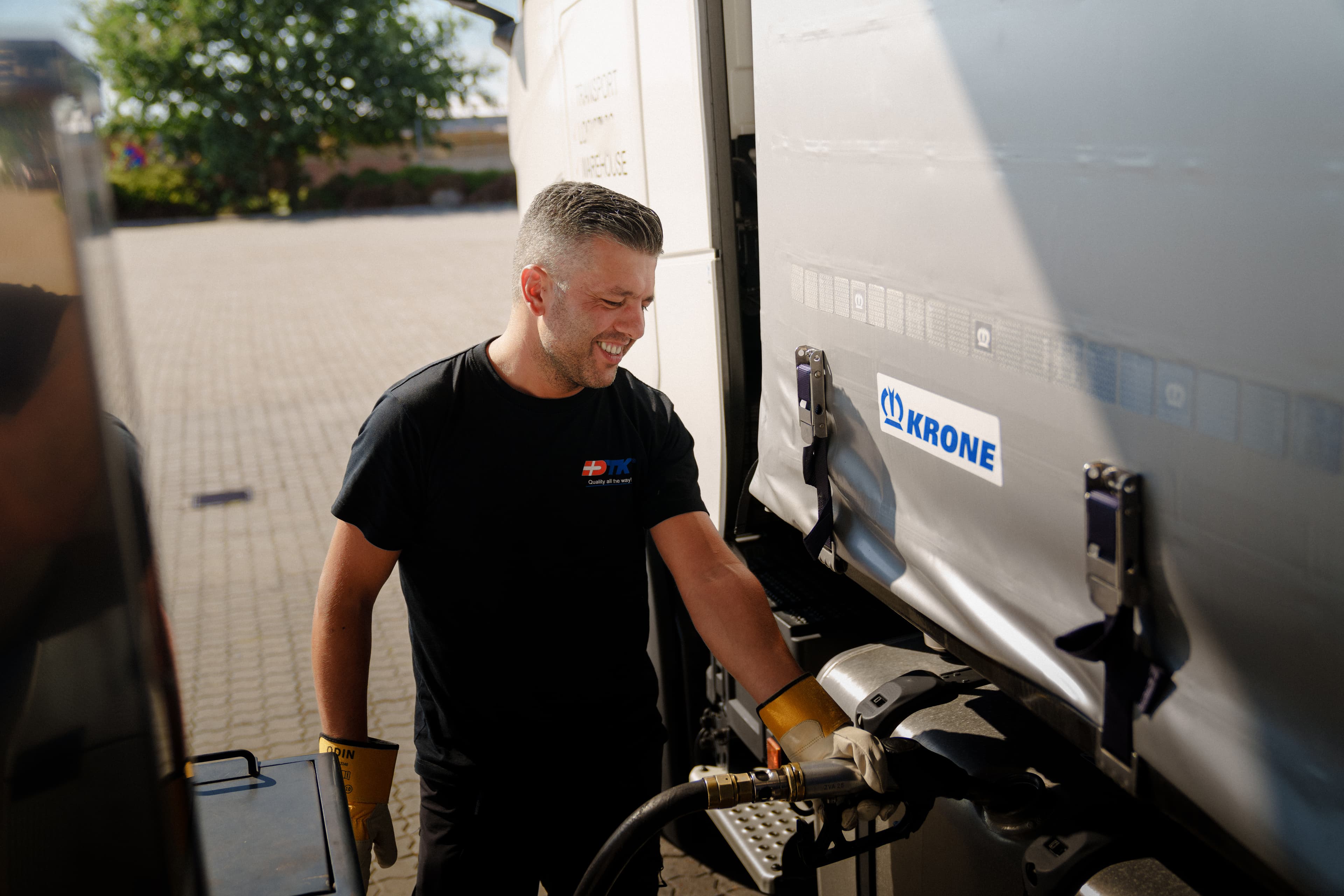 Smilende chauffør fylder diesel på sin lastbil ved Uno-X station, iført handsker og sort t-shirt, på en solrig dag.